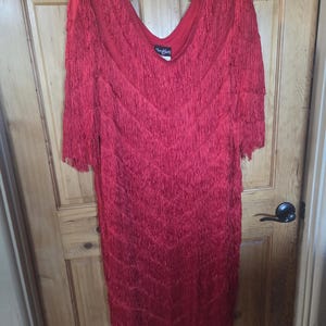 May include: A bright red knee-length dress featuring multiple horizontal layers of fringe covering the entire garment. The dress has a V-neckline and three-quarter length sleeves. A black label with the white text "Tee Chez" is visible.