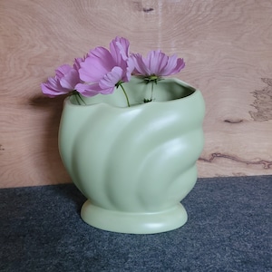 May include: A light green ceramic vase with a wavy design, holding pink cosmos flowers.