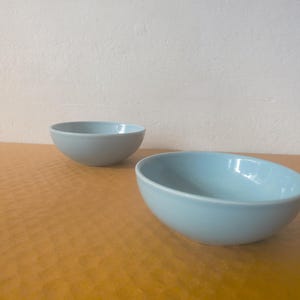 May include: Two smooth, light blue ceramic bowls with rounded edges and a glossy finish. The bowls are simple in design, featuring a consistent pale blue color throughout the interior and exterior surfaces, presented in a clean, minimalist style.