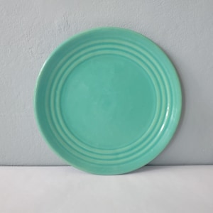 May include: A round, turquoise plate with three concentric rings near the rim. The plate has a smooth, glossy finish and is set against a light gray background. This plate is suitable for serving food.