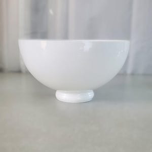 May include: A solid white ceramic bowl with a smooth, glossy finish and a rounded, deep basin. The bowl features a distinct, raised circular pedestal base that elevates the main body, providing a stable and balanced structure for the vessel.