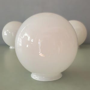 May include: Three white globe light fixtures with a glossy finish. The spherical shades sit on a small, circular base. The background is a muted gray-green.