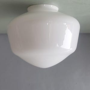 May include: A white glass light fitting with a rounded top and a flared, downward-tapering body. The fitting is designed for ceiling mounting. The smooth surface reflects light, creating a soft glow.