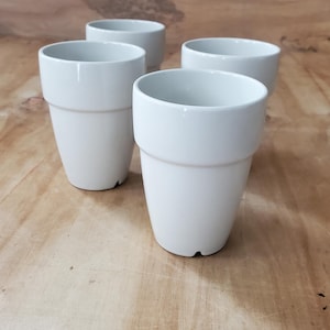 May include: A set of four white ceramic cups with a tapered base and a distinct rimmed top section. Each cup features a smooth, glossy finish and a simple, functional design, arranged in a cluster to display their uniform shape and color.