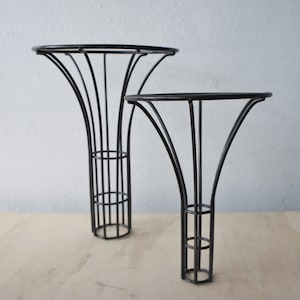 May include: Two black metal plant stands. The stands have a flared, trumpet-like shape with a circular top and a cage-like base. The stands are of different heights, with one taller than the other. The stands are on a light wood surface.