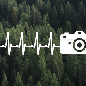 May include: A white camera icon with a heartbeat line in front of it, set against a background of green pine trees.