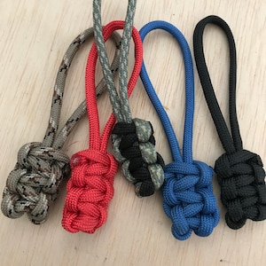 Puede incluir: Cinco tiradores de cremallera de diferentes colores con un nudo en el extremo. Los colores son camuflaje, rojo, negro y verde, azul y negro.