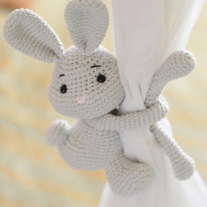 Puede incluir: Un juguete de conejo de crochet gris agarrado a una cortina blanca. El conejo tiene ojos negros y una nariz rosa.
