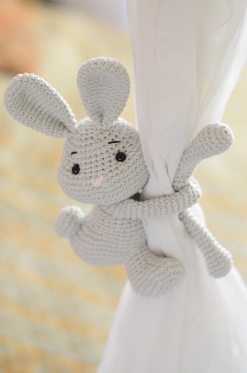 Puede incluir: Un juguete de conejo de crochet gris agarrado a una cortina blanca. El conejo tiene ojos negros y una nariz rosa.