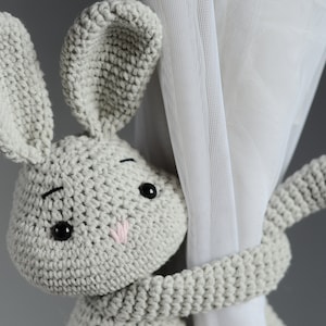 Puede incluir: Un juguete de conejo de crochet gris que sostiene una cortina blanca. El conejo tiene ojos negros y una nariz rosa.