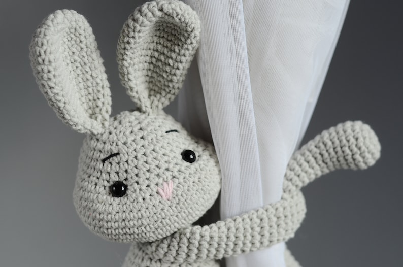 Puede incluir: Un juguete de conejo de crochet gris que sostiene una cortina blanca. El conejo tiene ojos negros y una nariz rosa.