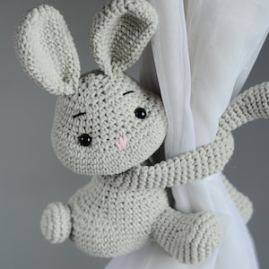 Puede incluir: Un juguete de conejo de crochet gris con ojos negros y una nariz rosa est&aacute; sujetando una cortina blanca. El conejo se utiliza como sujeta cortinas.