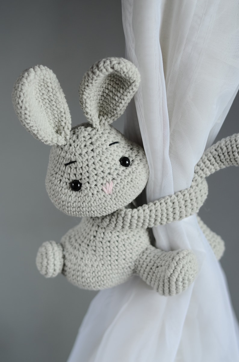 Puede incluir: Un juguete de conejo de crochet gris con ojos negros y una nariz rosa est&aacute; sujetando una cortina blanca. El conejo se utiliza como sujeta cortinas.
