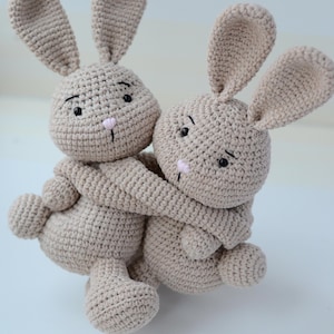 Puede incluir: Dos peluches de conejo de punto beige abraz&aacute;ndose. Los conejos tienen ojos negros y narices rosas.