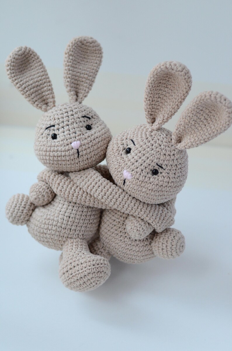 Puede incluir: Dos peluches de conejo de punto beige abraz&aacute;ndose. Los conejos tienen ojos negros y narices rosas.