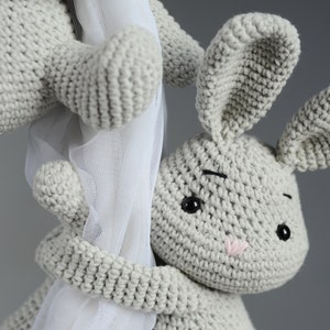 Puede incluir: Un juguete de conejo de crochet gris con ojos negros y una nariz rosa. El conejo se est&aacute; agarrando a una cortina blanca transparente.