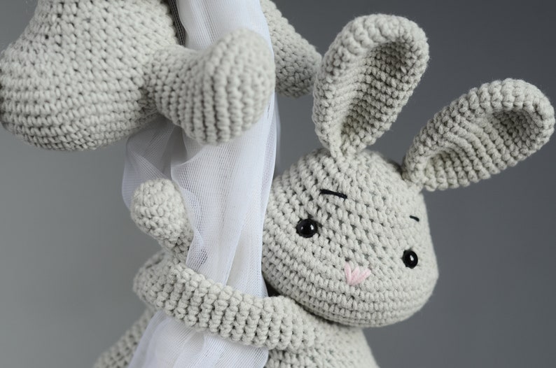 Puede incluir: Un juguete de conejo de crochet gris con ojos negros y una nariz rosa. El conejo se est&aacute; agarrando a una cortina blanca transparente.