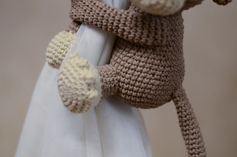 May include: A brown crocheted monkey toy with a cream-colored face and hands, holding onto a white curtain.