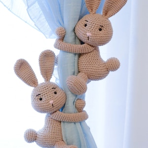 Puede incluir: Dos lazos de cortina de conejo de crochet de color marr&oacute;n claro sujetando una cortina de voile azul claro.