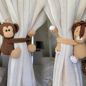 May include: Two crocheted stuffed animals, a brown monkey and a beige lion, are used as curtain tiebacks. The monkey is holding onto the curtain with its arms and tail, while the lion is holding onto the curtain with its paws.