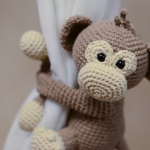 May include: A brown crocheted monkey toy with black eyes and a beige nose, holding onto a white curtain.