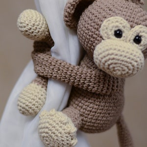 May include: A brown crocheted monkey toy with a cream-colored face and tail, holding onto a white curtain.