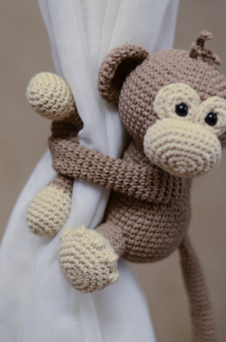 May include: A brown crocheted monkey toy with a cream-colored face and tail, holding onto a white curtain.