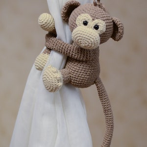 May include: A brown crocheted monkey toy with a long tail, holding onto a white curtain. The monkey has a beige face with black eyes.