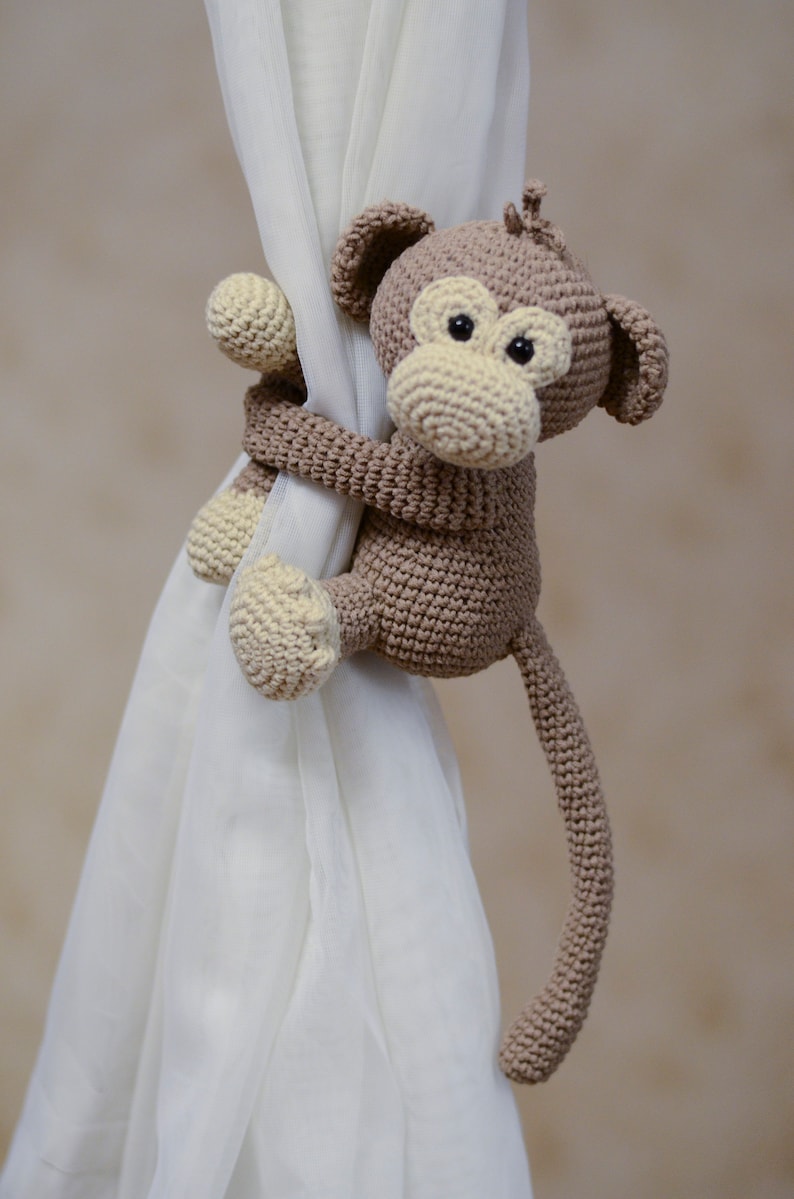 May include: A brown crocheted monkey toy with a long tail, holding onto a white curtain. The monkey has a beige face with black eyes.