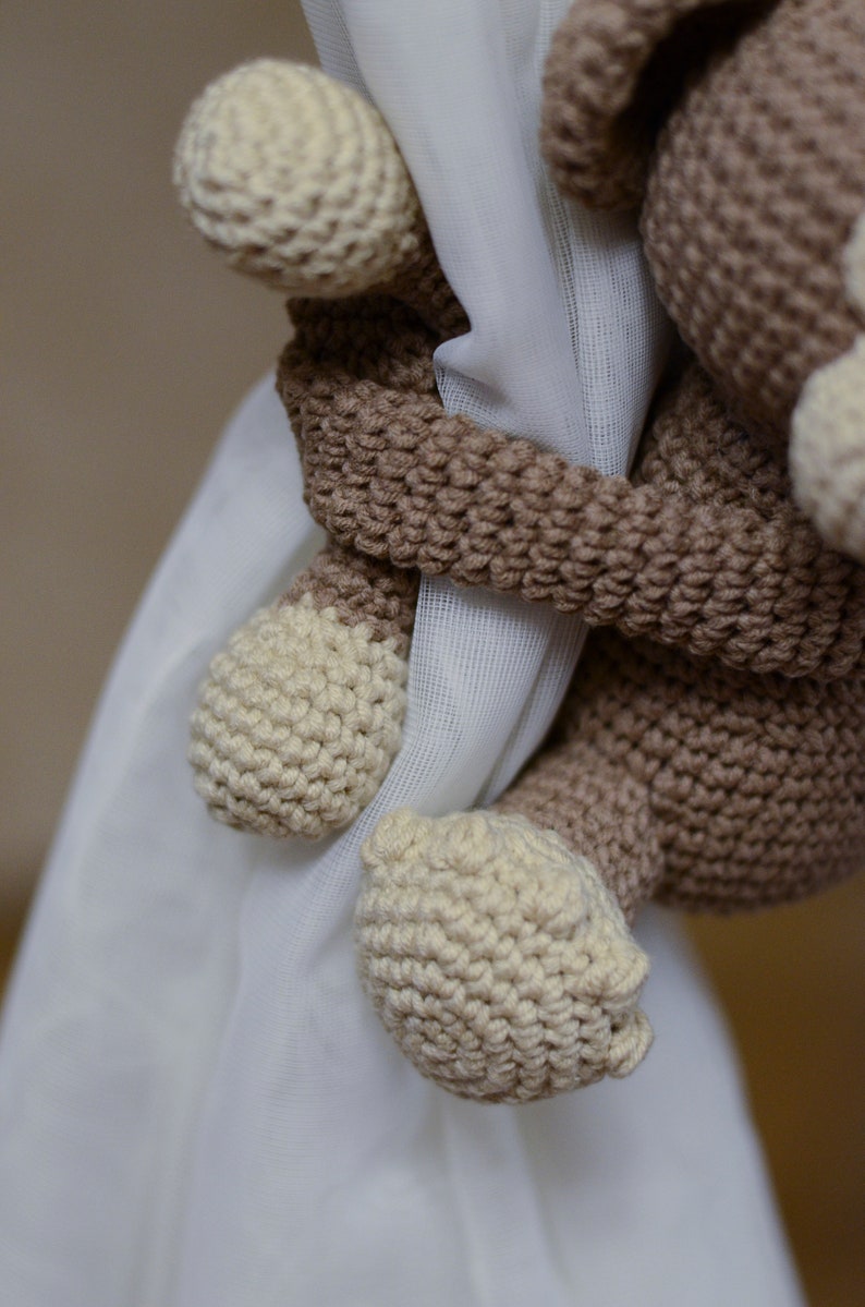 May include: A crocheted brown and cream monkey toy holding onto a white curtain. The monkey has a long tail and is holding onto the curtain with its hands.
