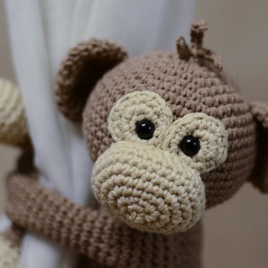 May include: A brown crocheted monkey toy with large black eyes and a beige nose. The monkey is holding onto a white curtain.