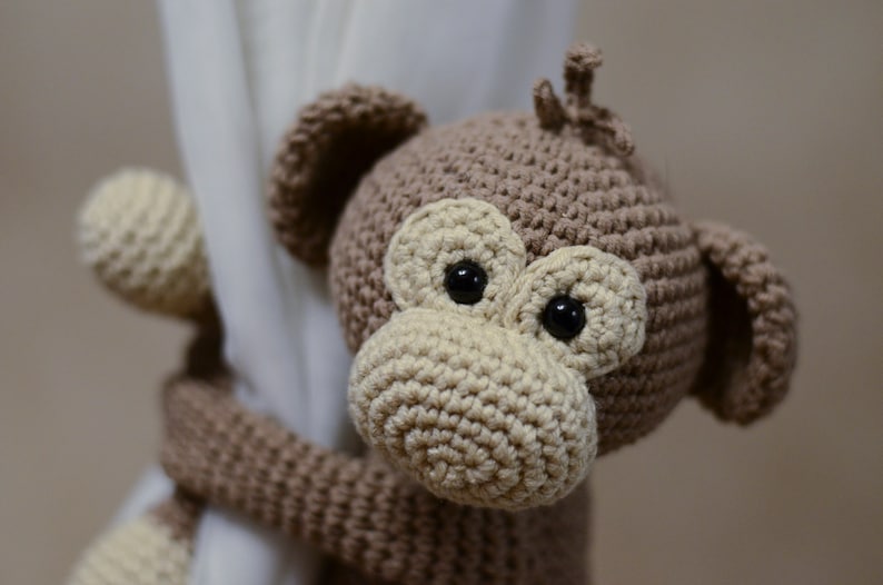 May include: A brown crocheted monkey toy with large black eyes and a beige nose. The monkey is holding onto a white curtain.