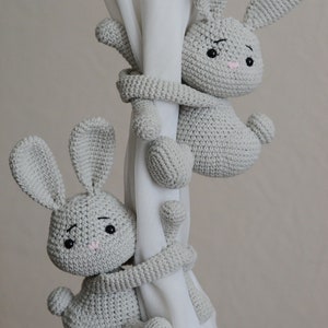 Puede incluir: Dos lazos de cortina de conejo de crochet gris. Los conejos est&aacute;n agarrando una cortina blanca con sus brazos y piernas.