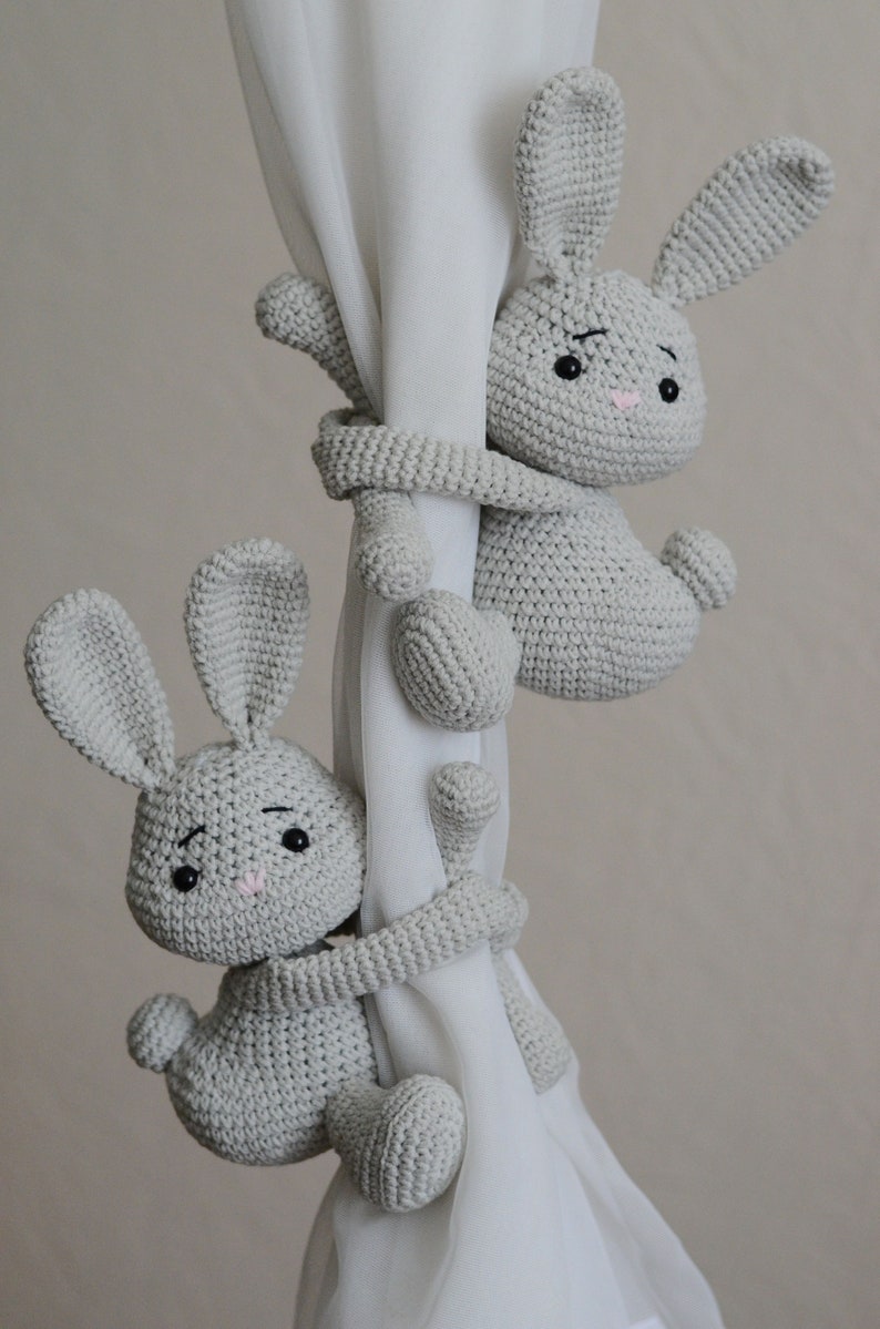 Puede incluir: Dos lazos de cortina de conejo de crochet gris. Los conejos est&aacute;n agarrando una cortina blanca con sus brazos y piernas.