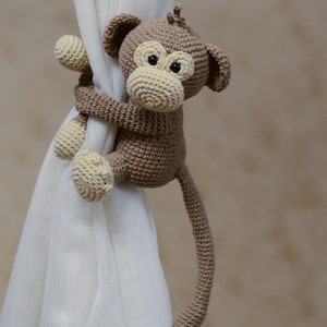 May include: A brown crocheted monkey toy with a long tail, holding onto a white curtain. The monkey has a brown body, a beige face, and black eyes.