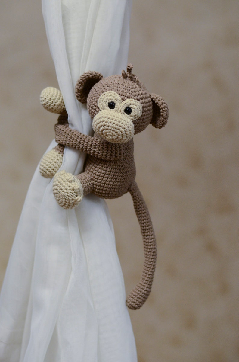 May include: A brown crocheted monkey toy with a long tail, holding onto a white curtain. The monkey has a brown body, a beige face, and black eyes.
