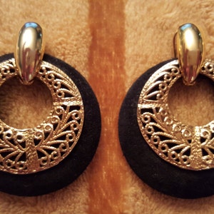 May include: A pair of gold and black earrings with a filigree design. The earrings are round and have a black velvet backing.