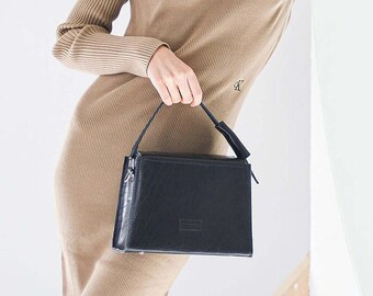 May include: A woman wearing a brown sweater dress and brown cowboy boots holds a black rectangular handbag. She is standing in front of a mirror.