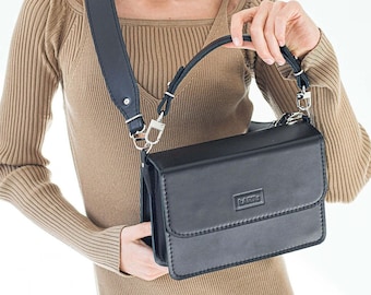 May include: A black leather crossbody bag with a silver buckle and a strap. The bag has a flap closure and a small logo on the front.