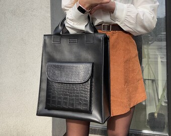 May include: A black leather tote bag with a crocodile embossed pocket. The bag has a top handle and a zipper closure.