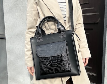 May include: A black leather tote bag with a crocodile embossed front pocket. The bag has a top handle and a long strap that can be worn over the shoulder.