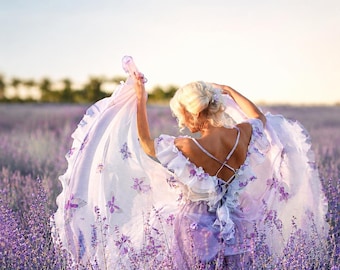 May include: A light purple dress featuring ruffled sleeves and a back with crisscross straps. The fabric is adorned with small purple floral appliqués and butterfly motifs, creating a layered, flowing skirt that is held up by the arms.
