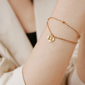 May include: A gold chain bracelet with a small circular pendant featuring a cross design. The bracelet has a delicate chain and small beads.