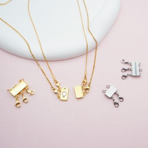 May include: Gold-tone and silver-tone magnetic necklace clasps with lobster claw clasps. The clasps are rectangular and have multiple attachment points for necklaces. The image shows the clasps with a gold chain necklace and on a pink surface.