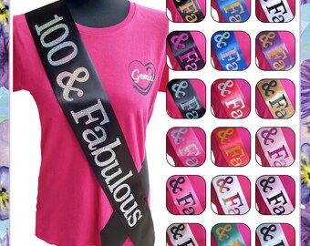 May include: A black satin sash with silver glitter lettering that reads "100 & Fabulous". The sash is worn over a pink t-shirt. The image also shows a variety of colors for the sash, including blue, purple, gold, red, pink, white, and teal.
