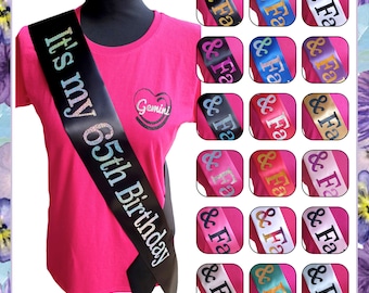 May include: A pink t-shirt with a black satin sash that says "It's my 65th Birthday". The sash is tied around the waist of the shirt. The shirt has a small glitter heart with the word "Gemini" inside. There are 16 different colored sashes in the background.
