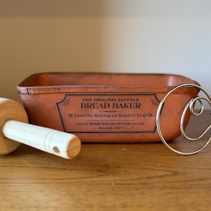 May include: A rectangular terracotta bread baker with a black label reading THE ORIGINAL SUFFOLK BREAD BAKER, Ideal for Baking an English Loaf, MADE BY HENRY WATSON POTTERY ENGLAND, Founded 1800, alongside a wooden rolling pin and a metal dough whisk.