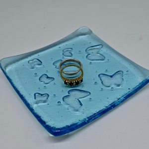 May include: A light blue glass ring dish with butterfly designs. The square dish has rounded corners and a raised edge. Two rings with gold bands and gemstones rest on the dish. The dish is approximately 10 cm square.