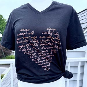 May include: Black V-neck t-shirt with a heart-shaped design in pink script. The design features dance terms like "stomp," "waltz," and "shuffle." The shirt is knotted at the waist.
