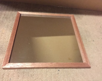 Peut inclure: Un petit miroir carré avec un cadre en bois. Le cadre est de couleur brun clair et a une finition lisse.